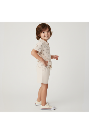 A child stands sideways, wearing a beige organic cotton collar tee with a playful print and matching shorts. The outfit is stylish and perfect for sunny day adventures.