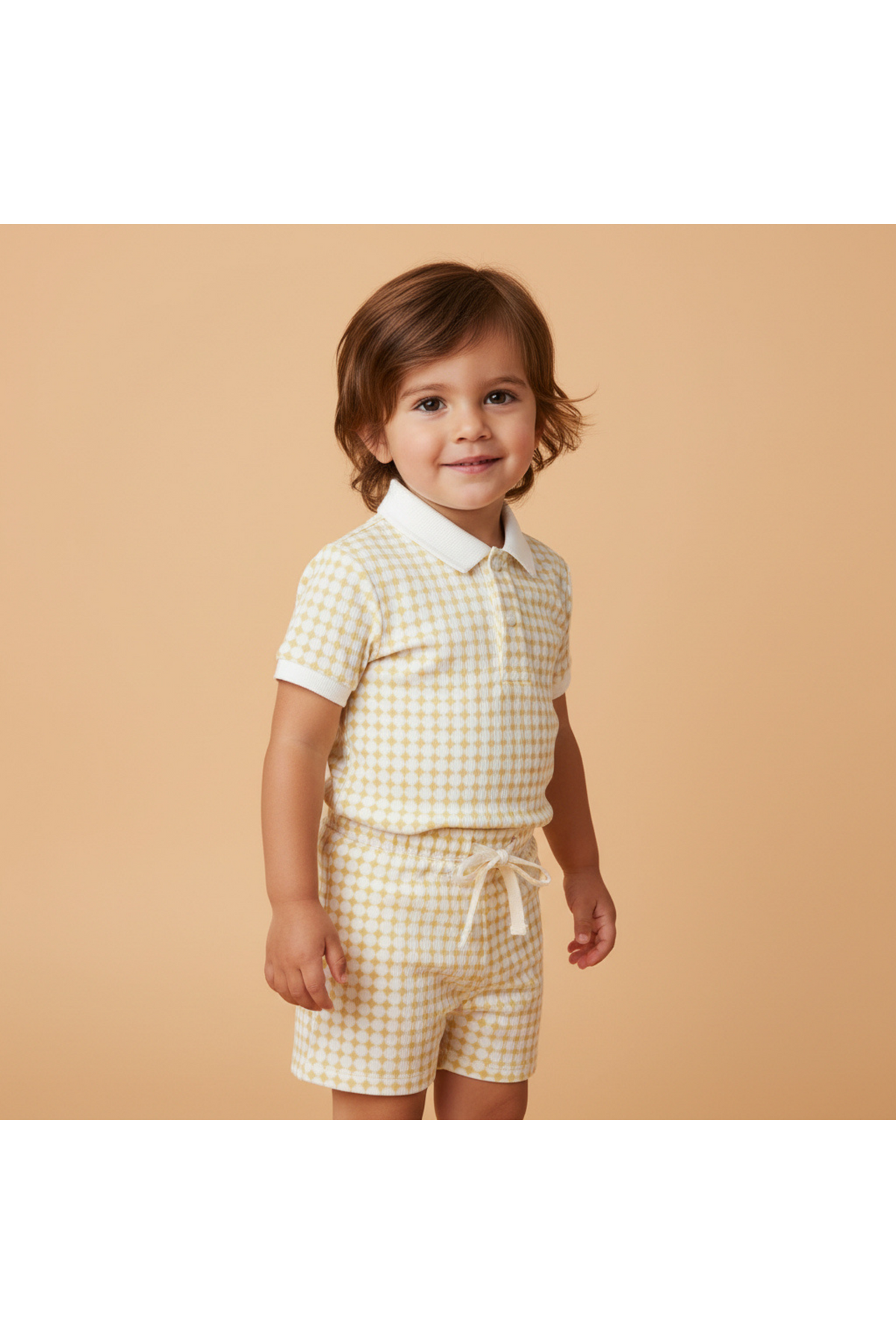 A smiling child wears a yellow and white gingham collar tee and shorts set, made from soft organic cotton. The background is a warm, neutral color, creating a cheerful atmosphere.