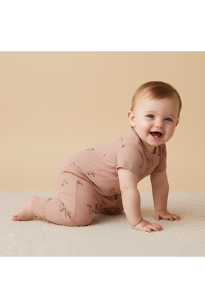 A smiling baby crawls on a soft, textured surface, wearing a light pink organic cotton romper with a subtle palm pattern.