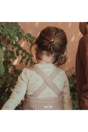 A young child with a ponytail wears a soft mauve knit overall over a light floral long-sleeve shirt, standing amidst greenery against a warm, textured background.