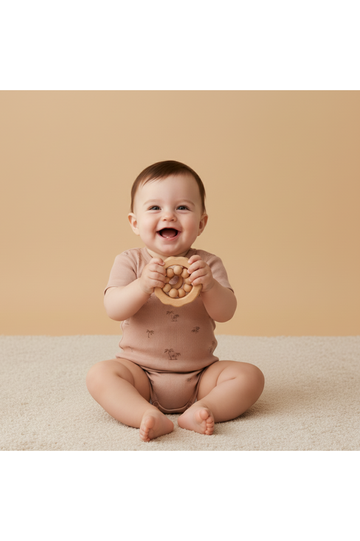 A smiling baby sits on a light-colored rug, wearing a soft, short-sleeve organic cotton romper in a muted pink shade. The baby holds a round wooden teething toy.