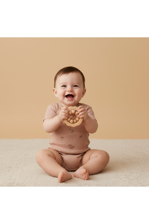 A smiling baby sits on a light-colored rug, wearing a soft, short-sleeve organic cotton romper in a muted pink shade. The baby holds a round wooden teething toy.
