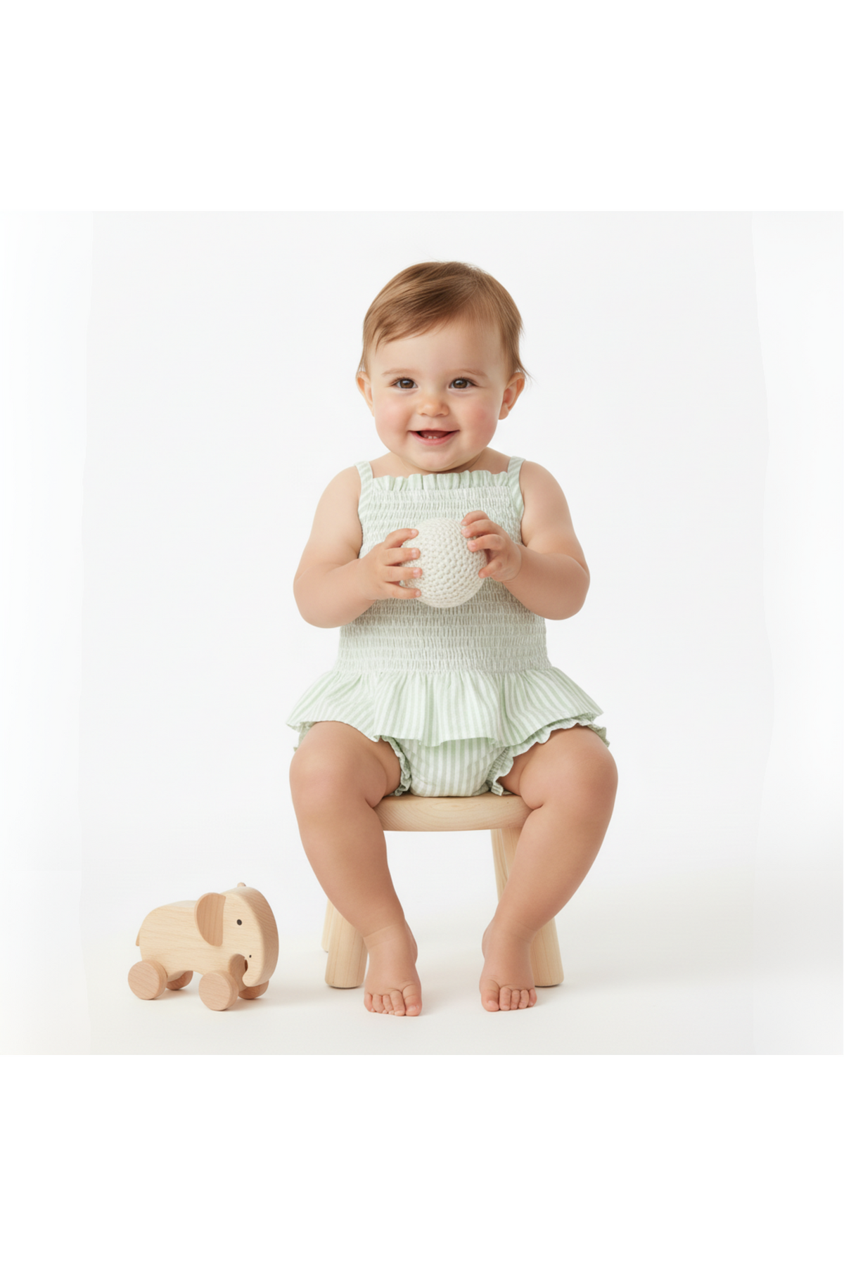 A smiling toddler sits on a wooden stool, wearing a light green smocked top and striped bloomer set.