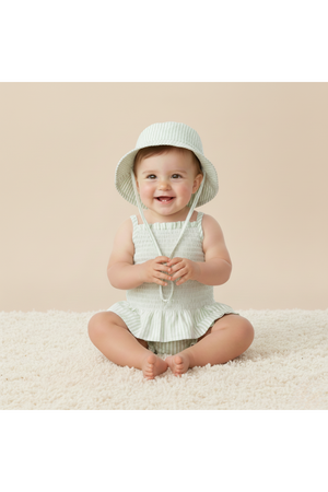 A smiling baby sits on a soft, textured surface, wearing a light green striped sun hat and matching outfit. The setting features a neutral beige background, highlighting the playful attire.