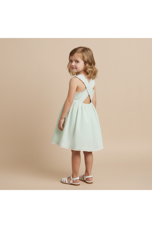 A young girl models a light green, organic cotton dress with criss-cross straps and a gathered skirt. The dress features a playful, breezy design perfect for warm weather.