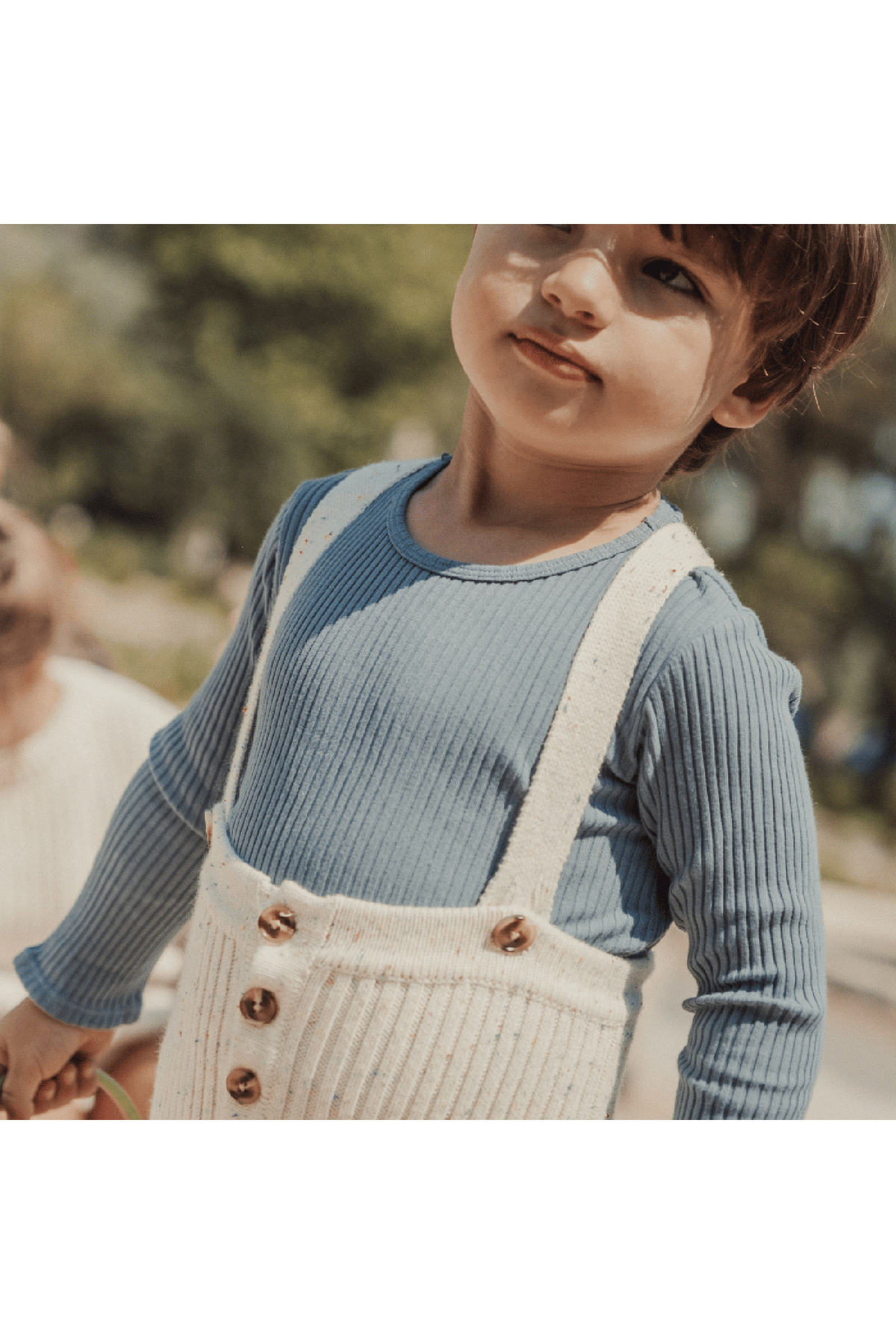A toddler wearing a blue ribbed long-sleeve shirt and cream-colored knit overall with wooden buttons stands outdoors, showcasing a playful and stylish outfit suitable for young children.