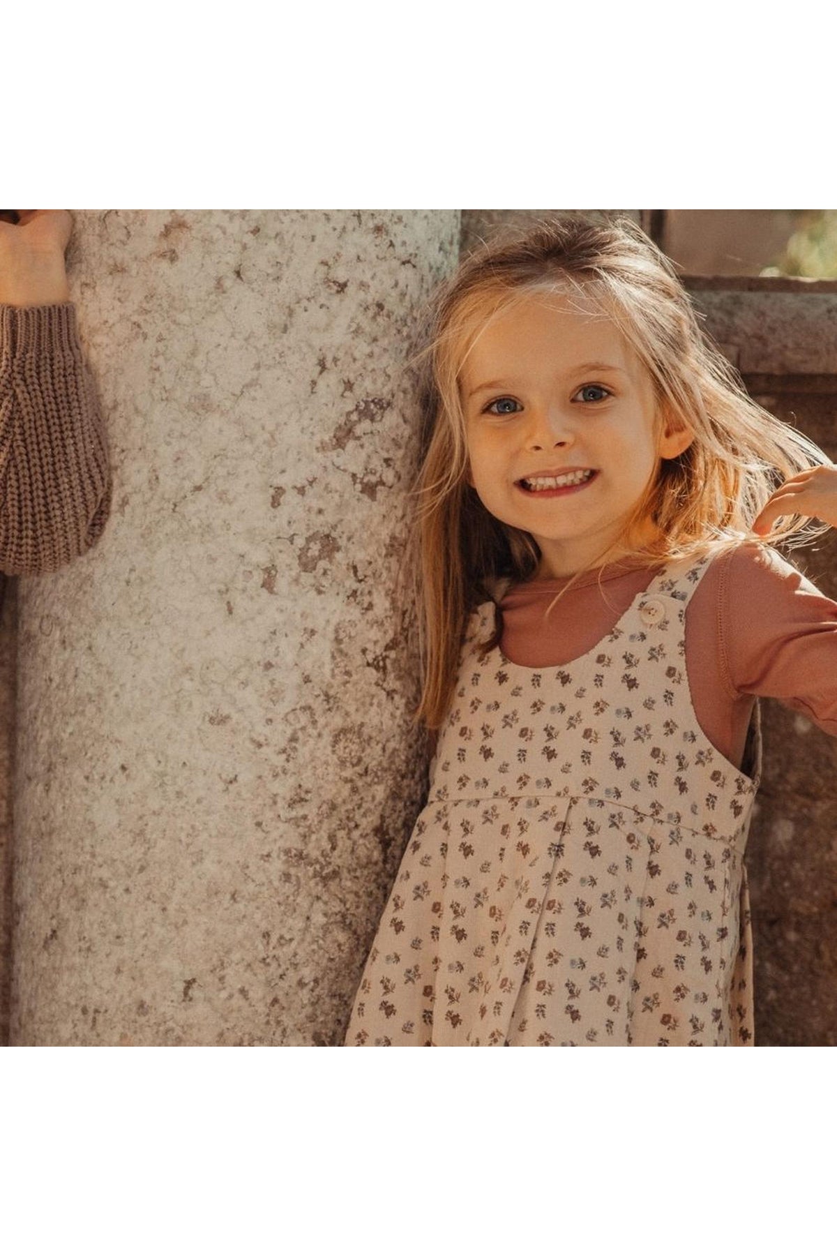 A smiling young girl stands beside a textured stone pillar, wearing a floral-print corduroy dress over a long-sleeve shirt. The dress features soft, muted colors, perfect for casual outings.