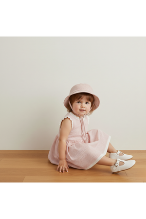 A toddler sits on a wooden floor, wearing a pink striped dress and a matching bucket hat.