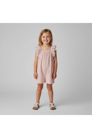 A smiling young girl is wearing a light pink, ruffled flutter jumpsuit. The outfit features a comfortable fit and is made from organic cotton, perfect for playful activities.