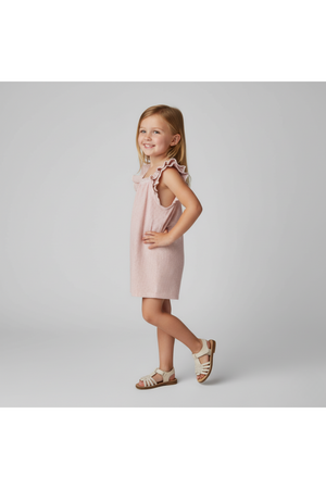 A smiling young girl stands in profile, wearing a light pink sleeveless dress with ruffle details. She pairs the outfit with white sandals, showcasing a playful and stylish look.