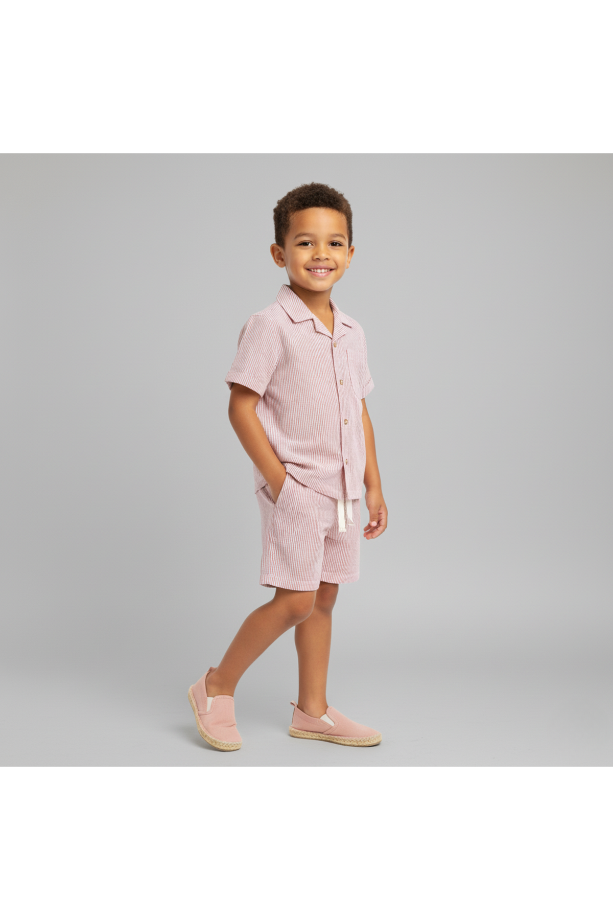 A young boy smiles while wearing a coral-striped organic shirt and shorts set. The lightweight fabric offers comfort and style, perfect for summer adventures.