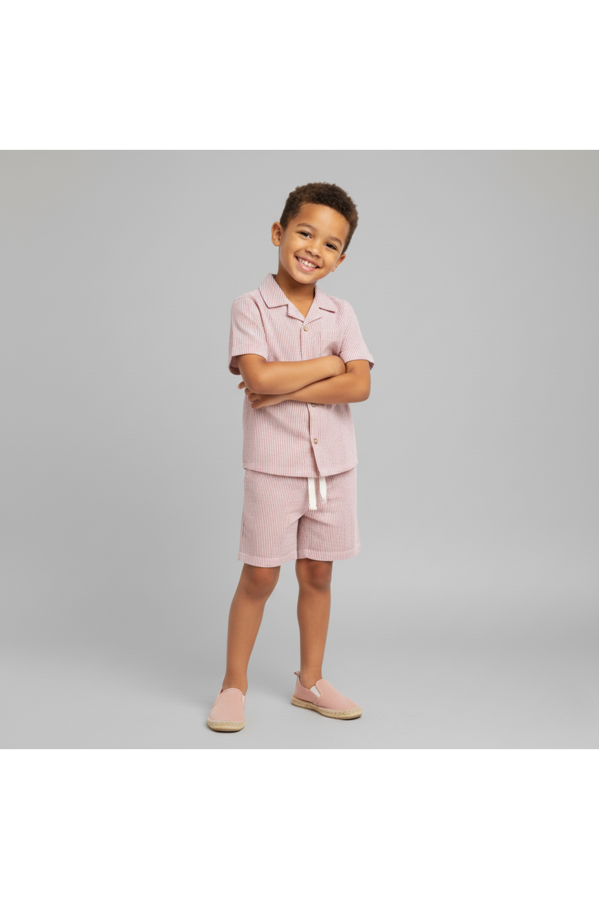 A young boy stands confidently, wearing a coral-striped organic cotton shirt and shorts set. The lightweight fabric is perfect for summer, showcasing a playful yet stylish design.