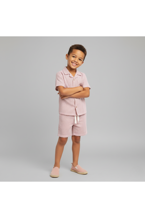 A young boy stands confidently, wearing a coral-striped organic cotton shirt and shorts set. The lightweight fabric is perfect for summer, showcasing a playful yet stylish design.