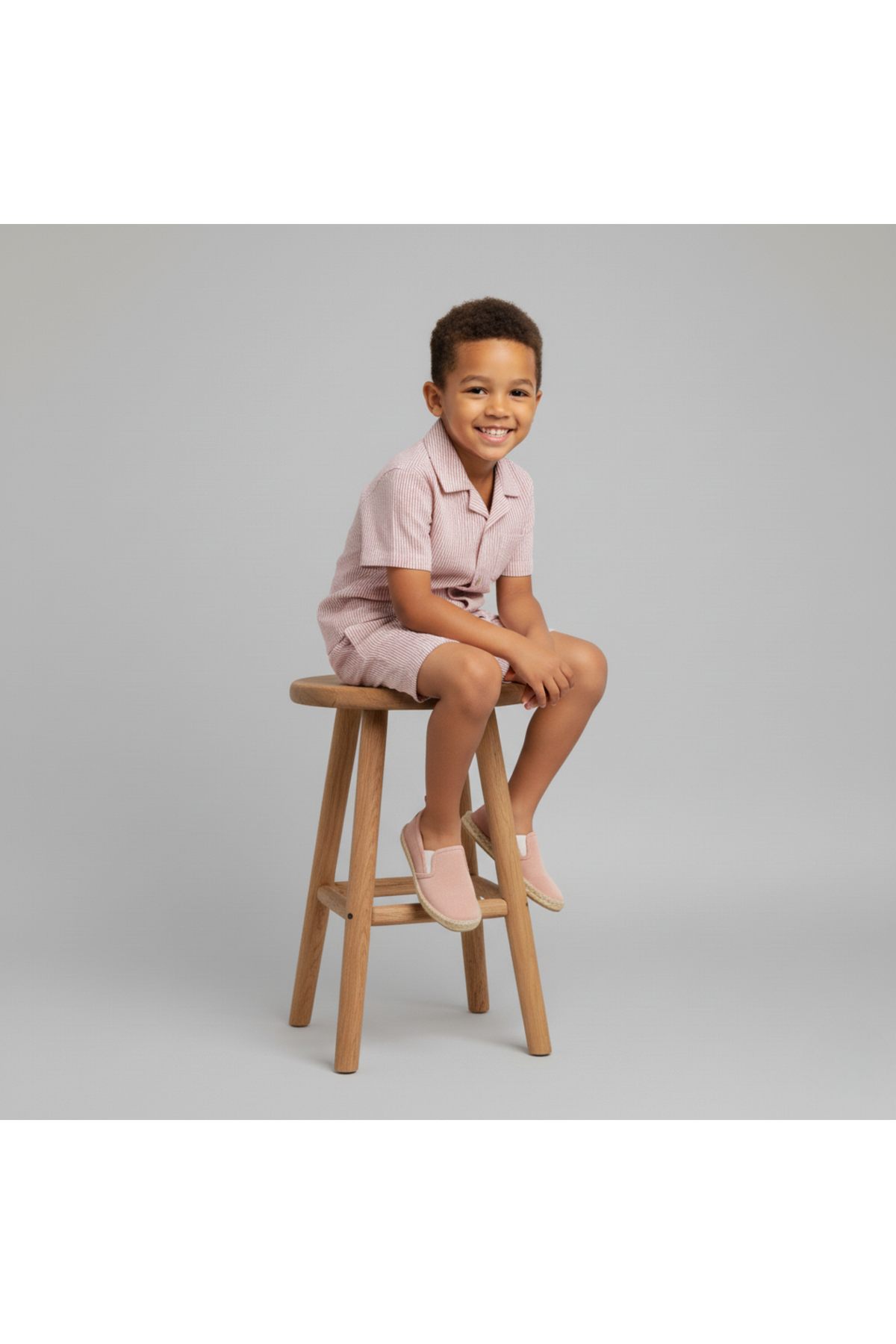 A young boy sits on a wooden stool, wearing a coral striped shirt and shorts set made from organic cotton. He smiles, showcasing a comfortable and stylish summer outfit.