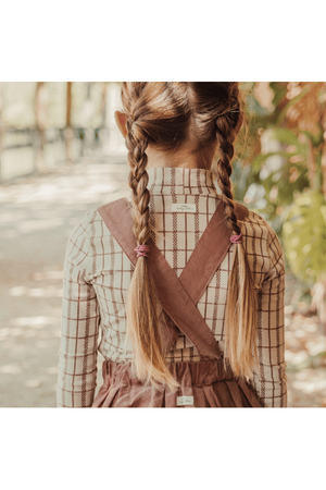 A young girl with braided hair wears a mauve check printed turtleneck and a brown pinafore dress, standing on a leaf-strewn path surrounded by greenery.
