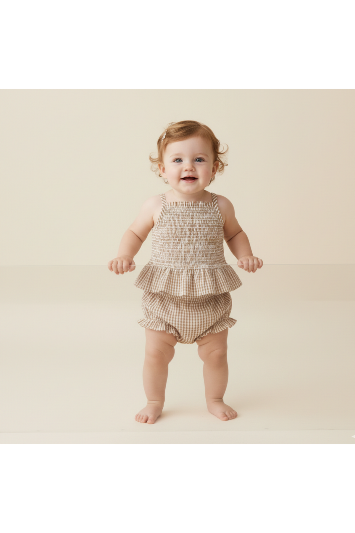 A smiling baby stands on a light background, wearing a caramel check organic smocked top and bloomer set.