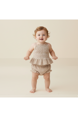 A smiling baby stands on a light background, wearing a caramel check organic smocked top and bloomer set.