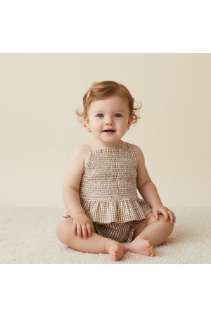 A smiling toddler sits on a soft surface, wearing a caramel check organic smocked top with delicate straps and matching bloomers.