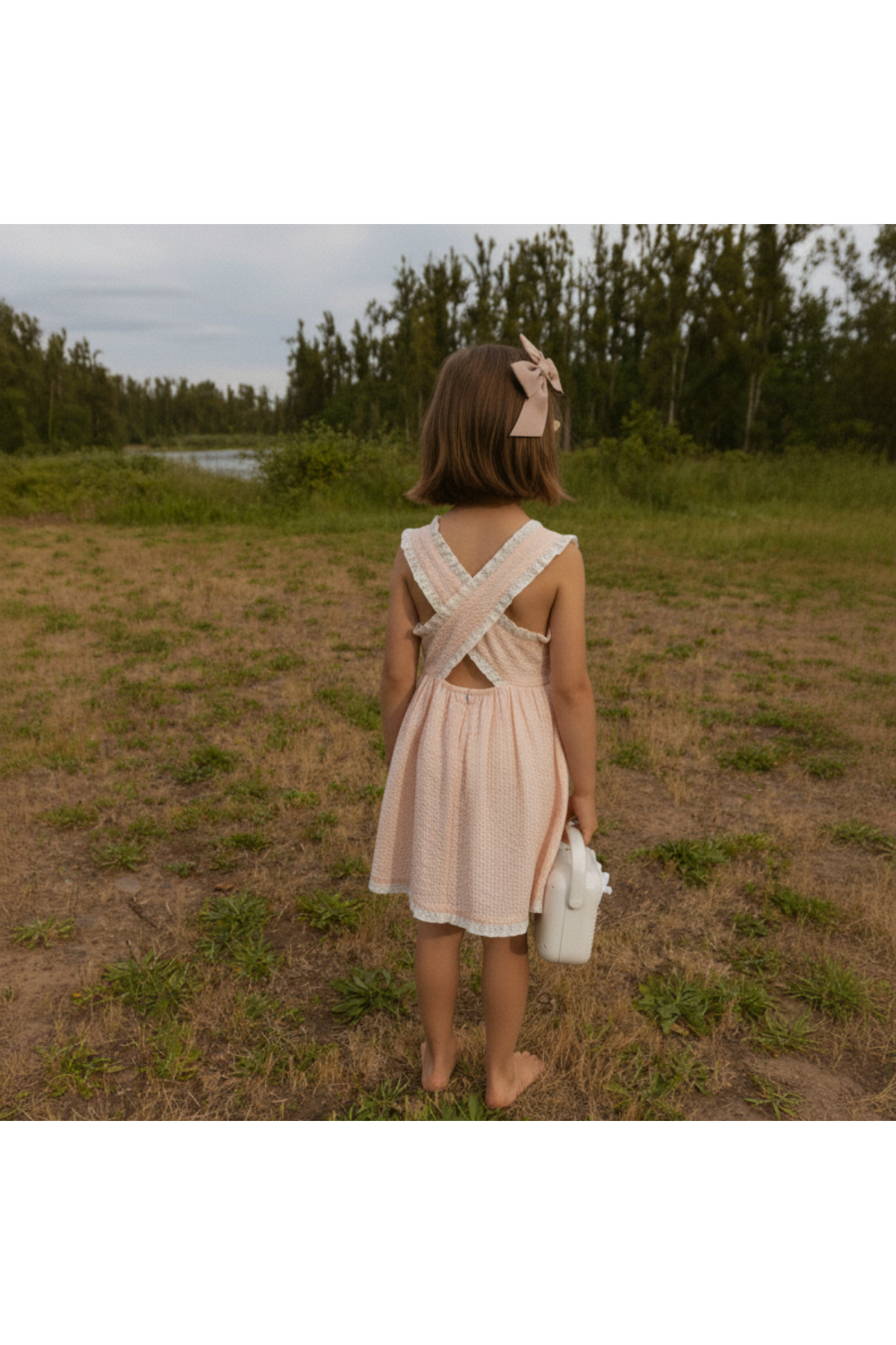 A young girl stands barefoot in a grassy field, wearing a blush-colored Organic Cross Back Dress with criss-cross straps and a gently gathered skirt, holding a small white container.