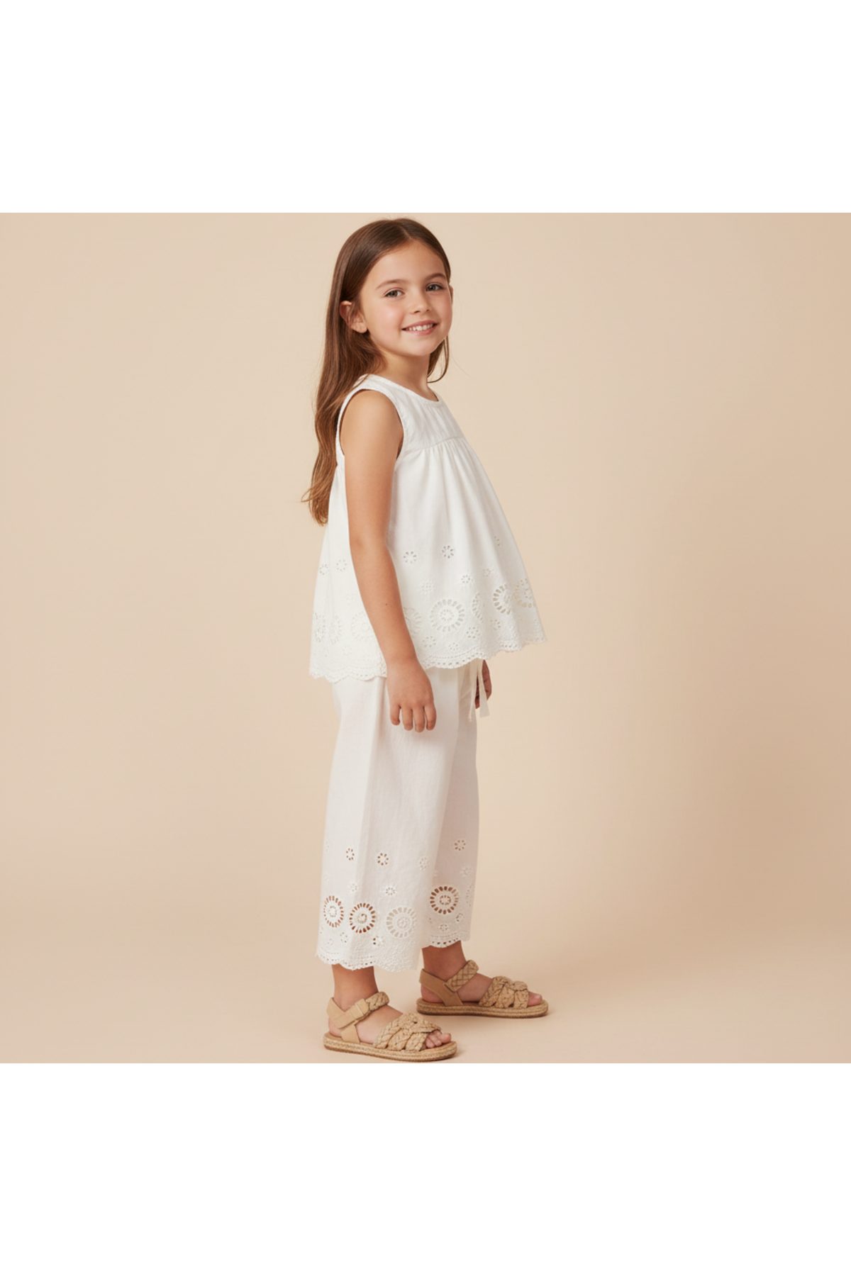 A young girl smiles while wearing an ivory organic cotton eyelet top and pant set. The outfit features intricate cutwork and a breezy design, perfect for warm-weather occasions.