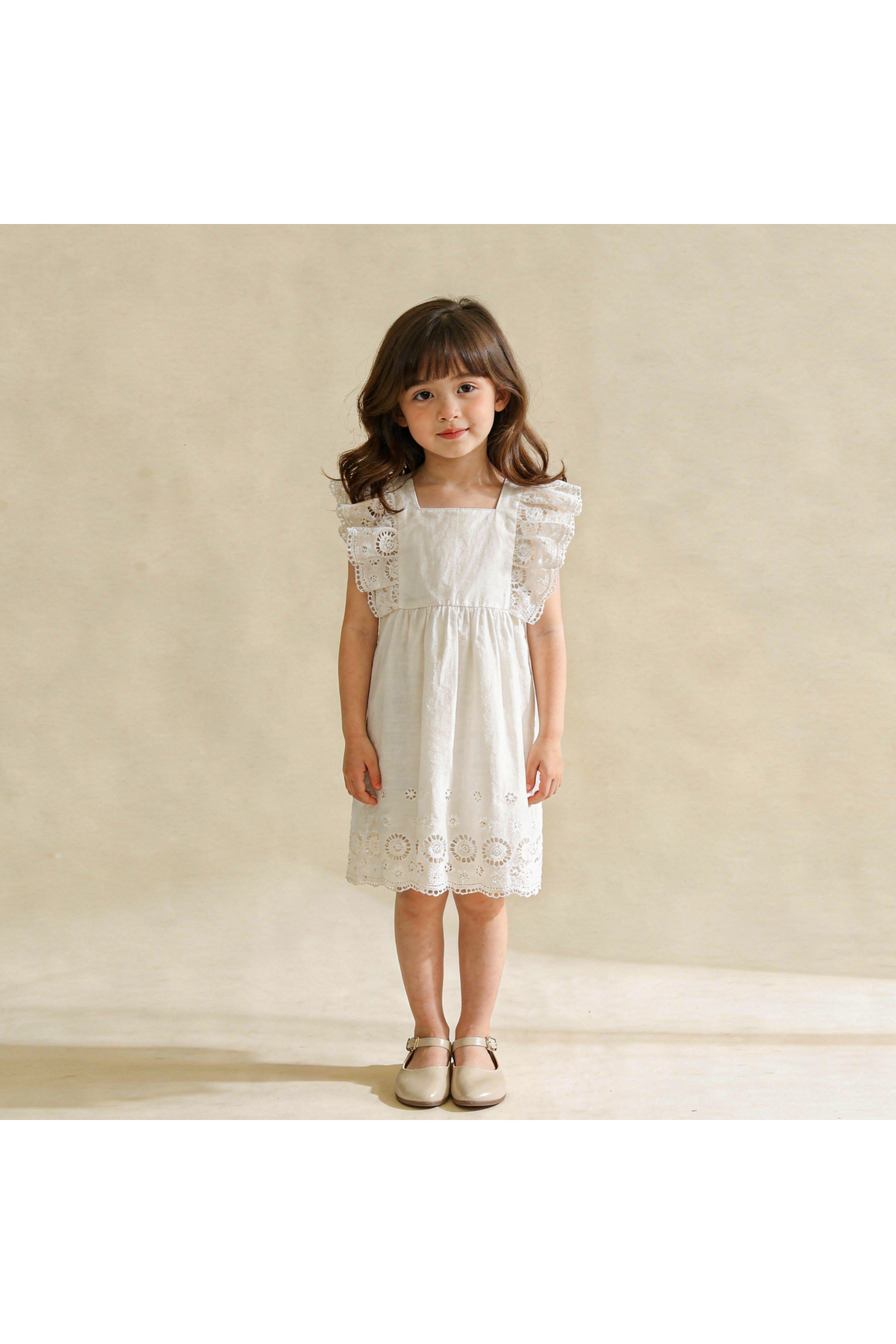 A young girl stands wearing an ivory organic cotton dress featuring delicate eyelet flutter sleeves and a lace hem. The dress is light and airy, perfect for playful occasions.