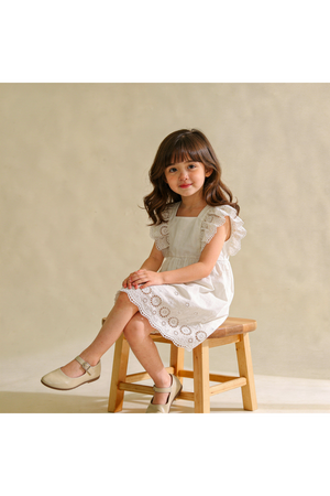 A young girl sits on a wooden stool, wearing a light ivory organic cotton dress with delicate flutter sleeves and eyelet detailing, showcasing a playful yet timeless style.