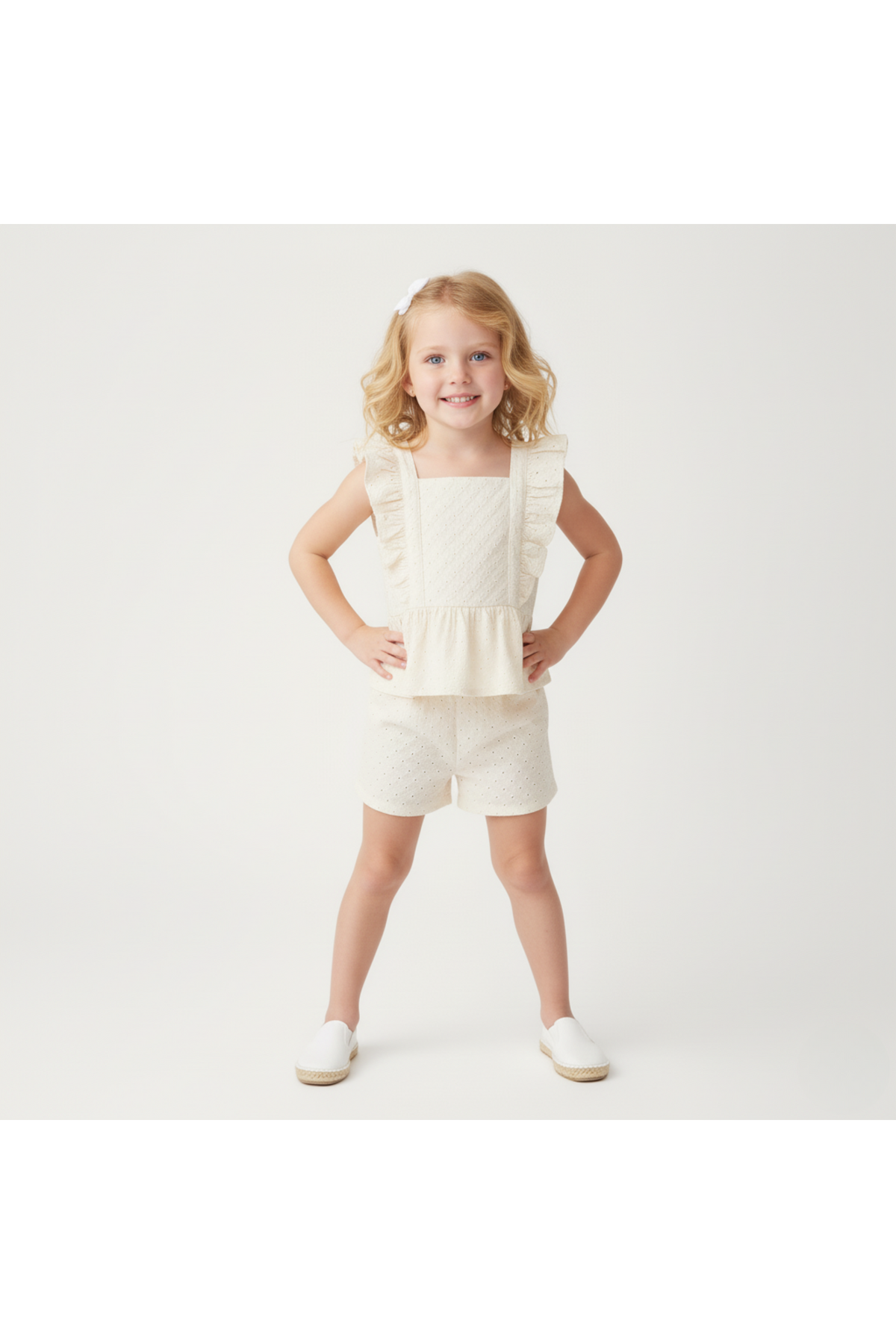 A young girl poses confidently in a light ivory organic cotton eyelet top with ruffled straps and matching shorts, showcasing a playful yet elegant style perfect for warm-weather outings.