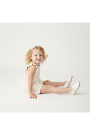 A smiling young girl sits on a light background, wearing a delicate ivory eyelet top with flutter sleeves and matching shorts, paired with white slip-on shoes.