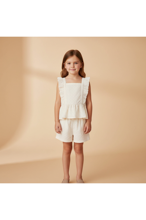 A young girl stands wearing a cream-colored organic cotton eyelet flutter top with ruffles and matching shorts.