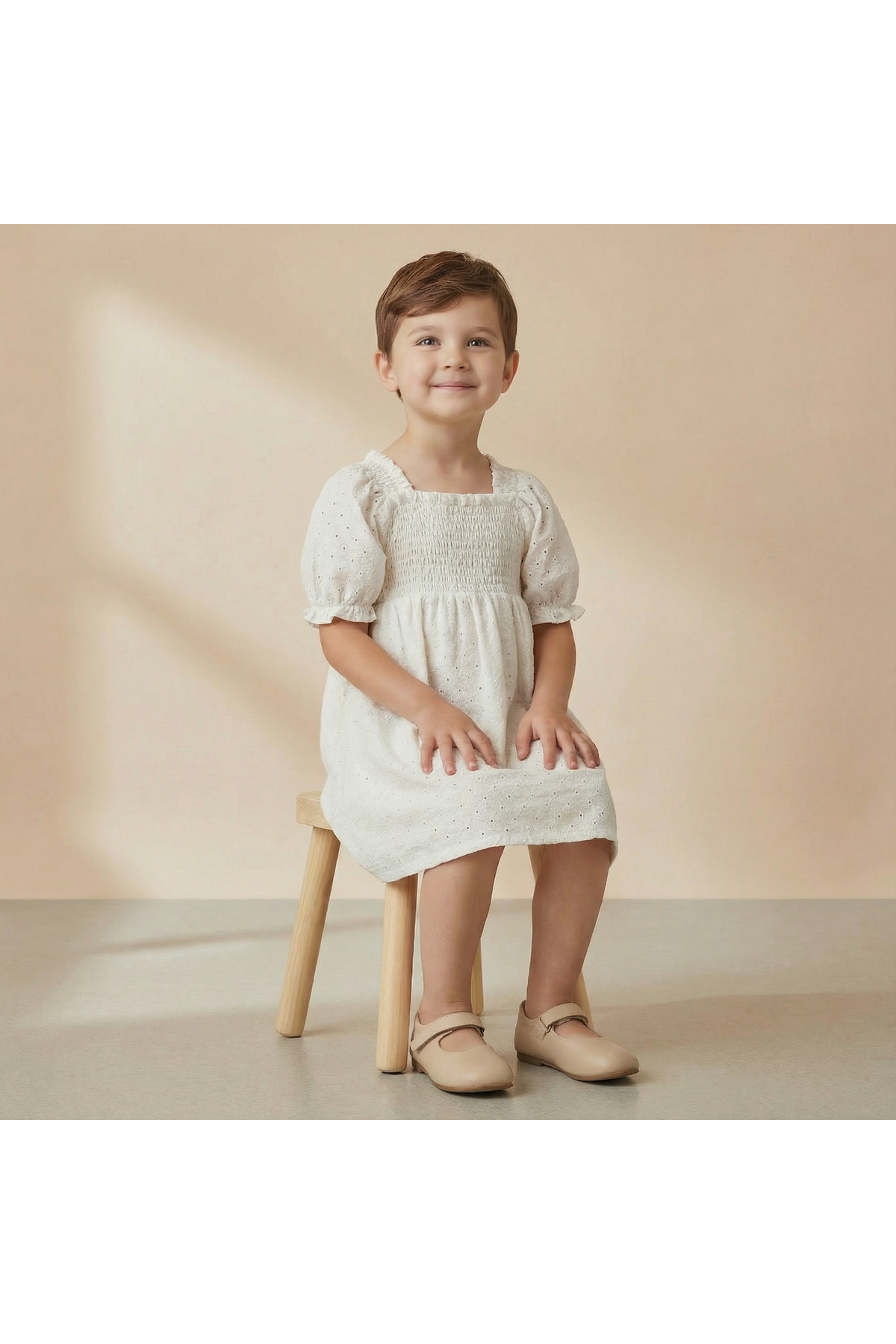 A young child sits on a wooden stool, wearing an ivory organic cotton eyelet bubble sleeve dress. The dress features delicate embroidery and a smocked bodice, perfect for warm weather.