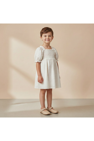A young child stands smiling in a soft, ivory eyelet bubble sleeve dress. The dress features delicate embroidery and a comfortable fit, perfect for warm-weather wear.