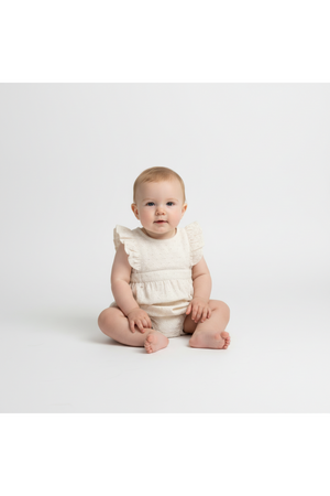 A seated baby wears an ivory eyelet romper with ruffled sleeves, showcasing delicate embroidery. The clean, neutral background emphasizes the organic cotton fabric and charming design for warm-weather outings.