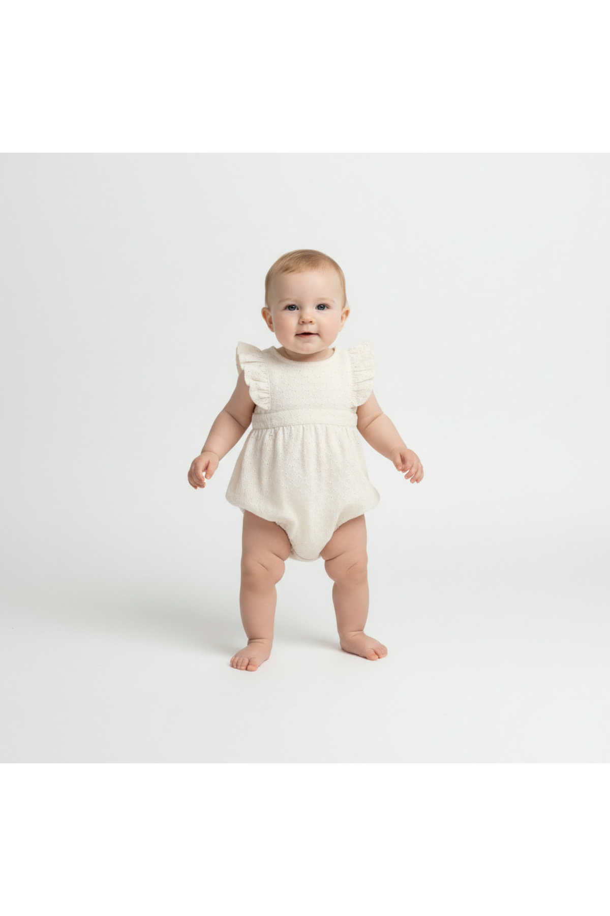 A smiling baby stands in a soft ivory organic cotton romper with delicate flutter sleeves. The outfit features a textured design, perfect for warm-weather outings and made for comfort.