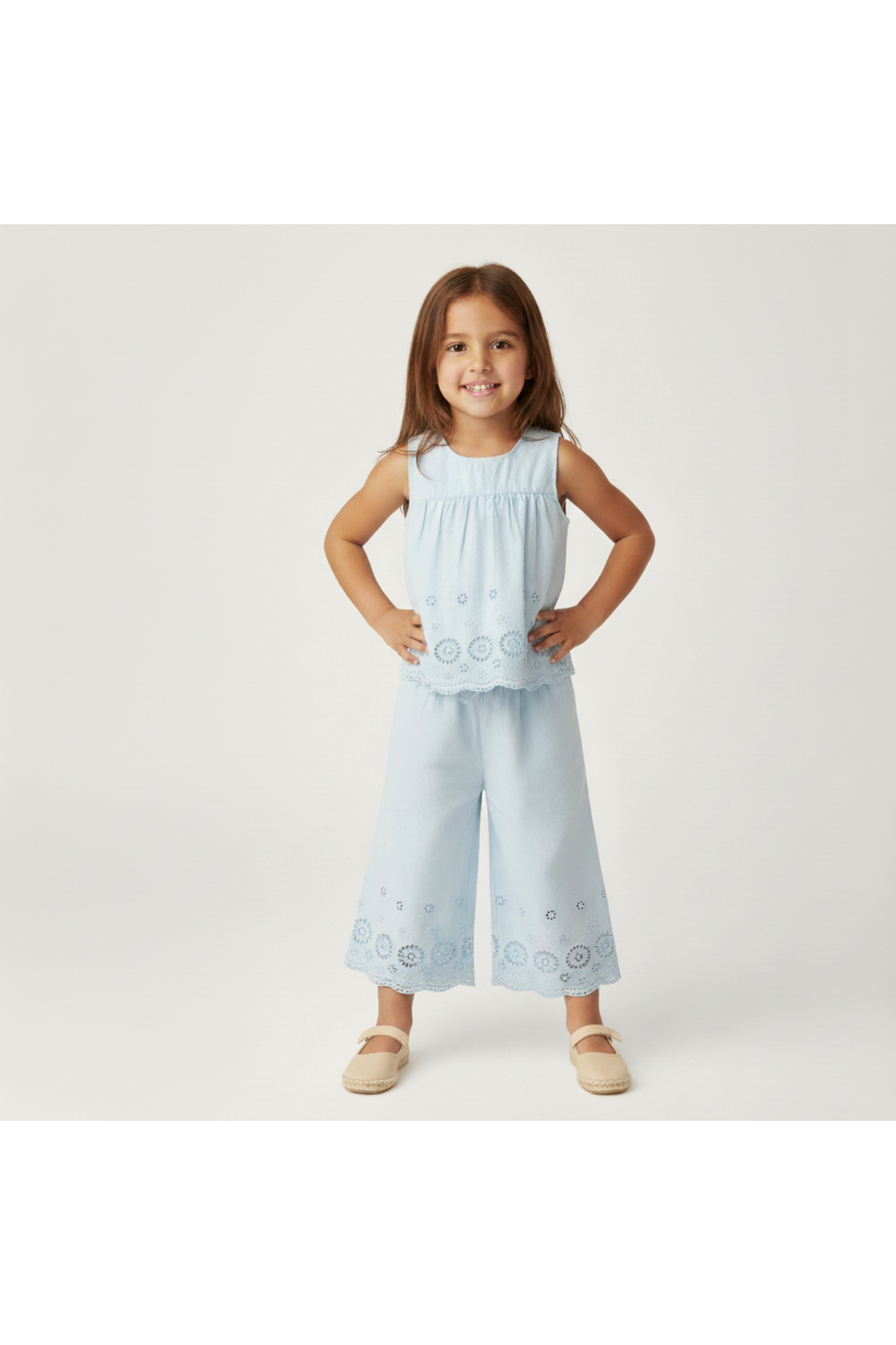 A young girl stands smiling in a light blue organic cotton eyelet top and matching pants set, featuring delicate embroidery and a breezy silhouette, perfect for warm-weather outings.