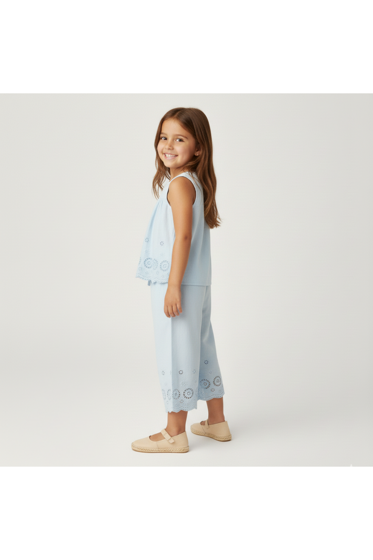 A young girl is smiling while wearing a light blue organic cotton eyelet top and matching pants set.