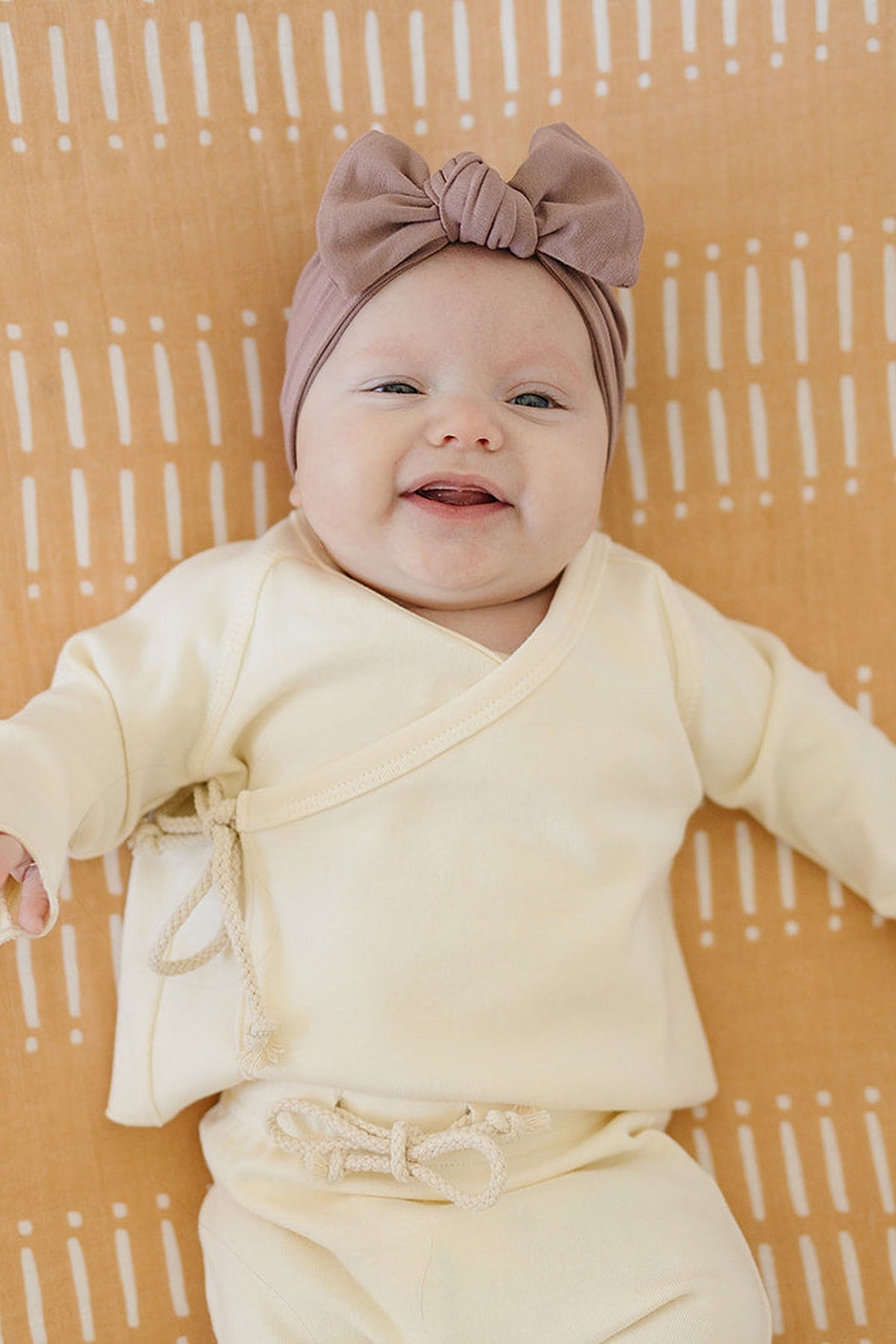 A baby wearing a dusty rose head wrap and cream outfit lies on a patterned orange blanket, smiling.