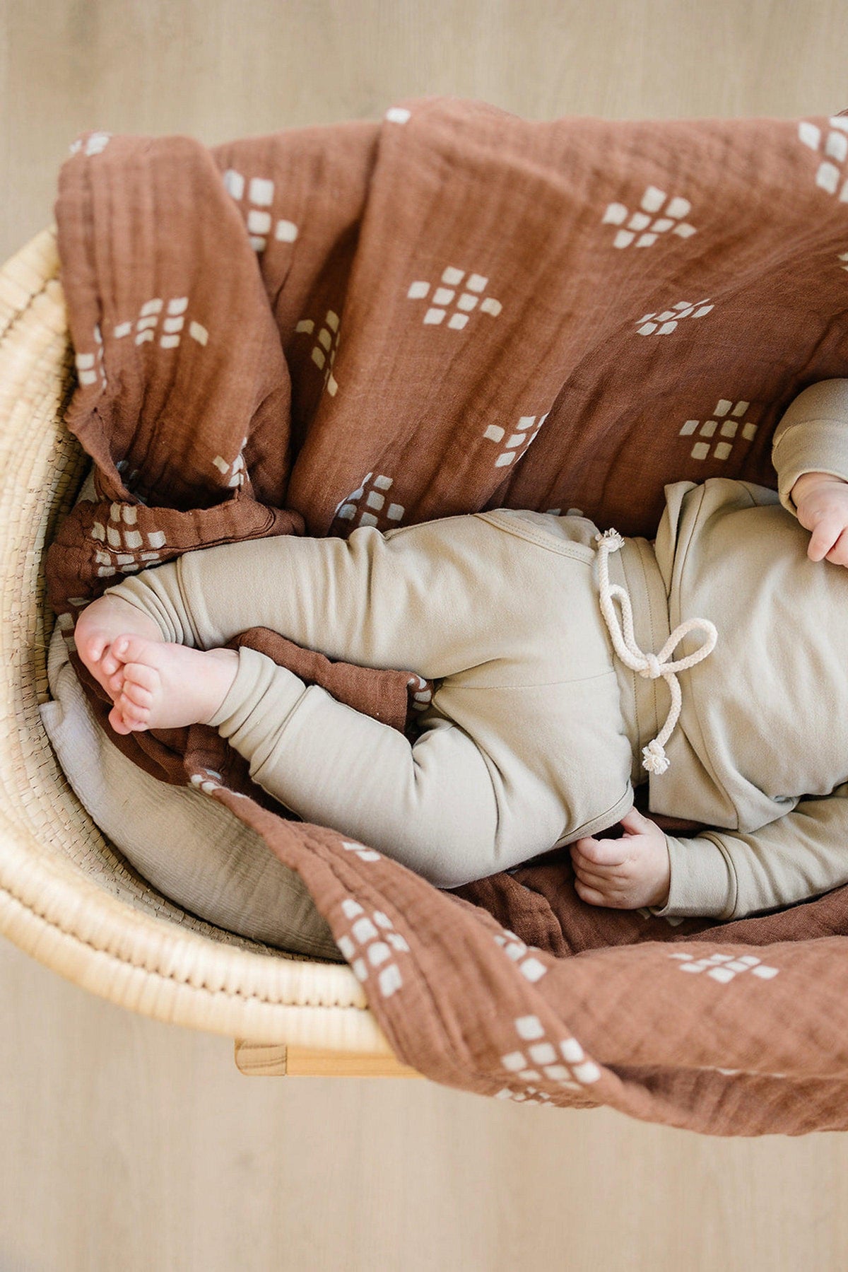 A baby is wrapped in a brown muslin quilt with white patterns, lying in a woven basket.