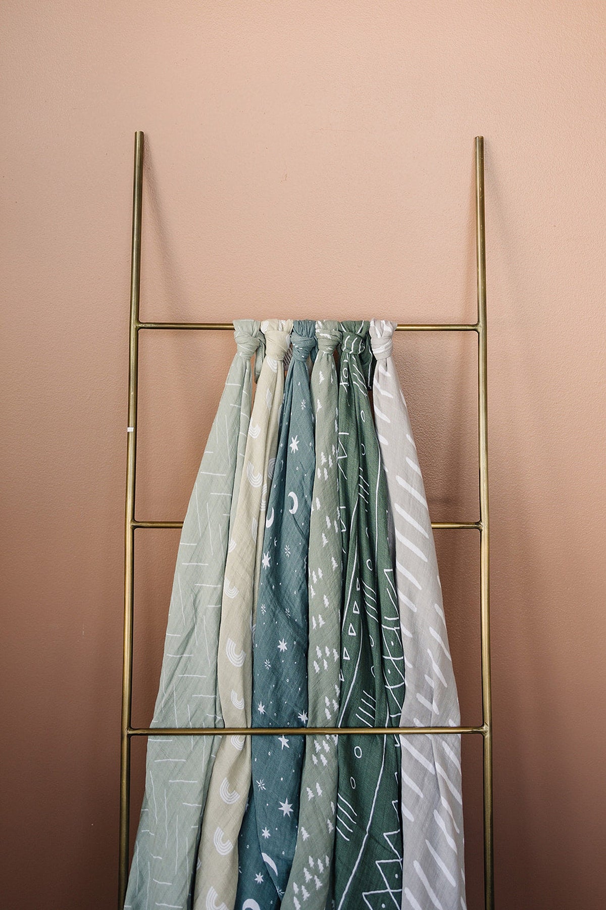 A set of patterned muslin swaddle blankets in various green tones hangs on a brass ladder against a peach wall.