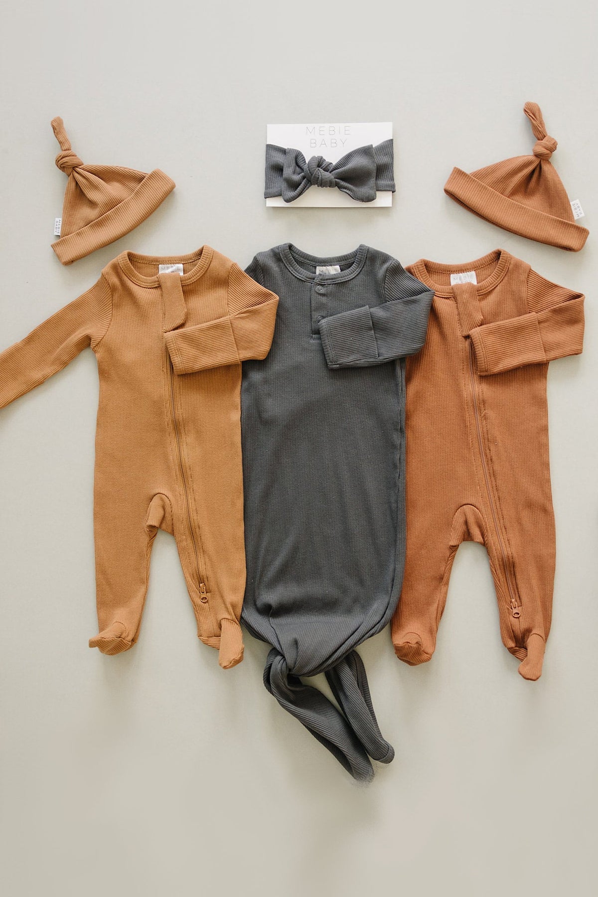 Three ribbed baby outfits in brown and charcoal with matching hats and a headband, displayed on a neutral background.
