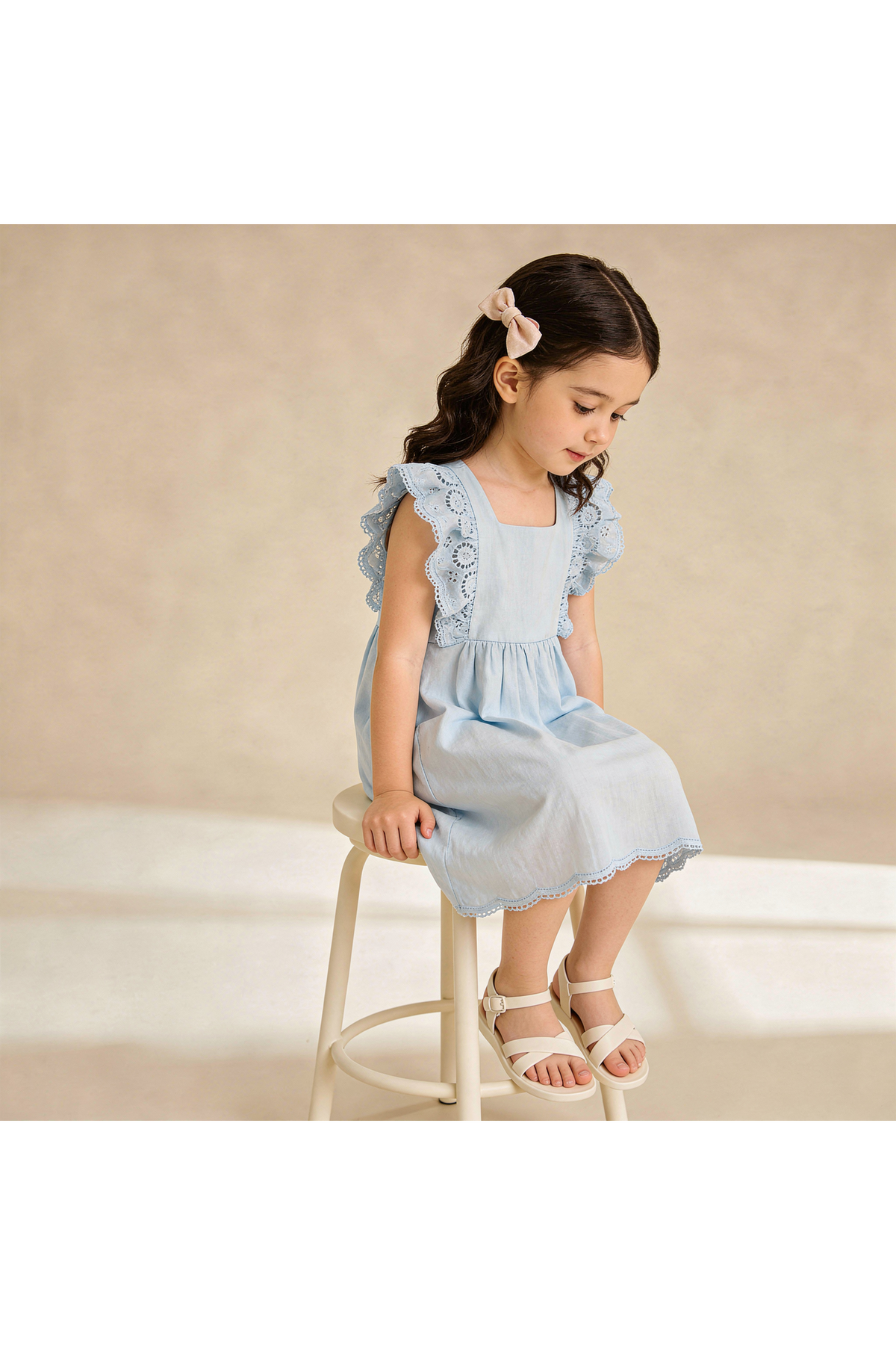 A young girl sits on a stool, wearing a light blue organic cotton dress with flutter sleeves and lace detailing. She has a hair accessory and a thoughtful expression.
