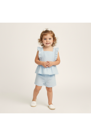 A smiling toddler wears a light blue organic cotton eyelet flutter top with ruffles and matching shorts. The outfit features delicate embroidery, perfect for warm-weather outings and celebrations.