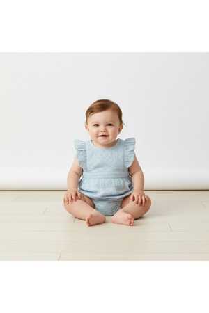 A smiling baby sits on a light-colored wooden floor, wearing a light blue organic cotton romper with delicate eyelet detailing and ruffled sleeves, showcasing a cute and comfortable style.
