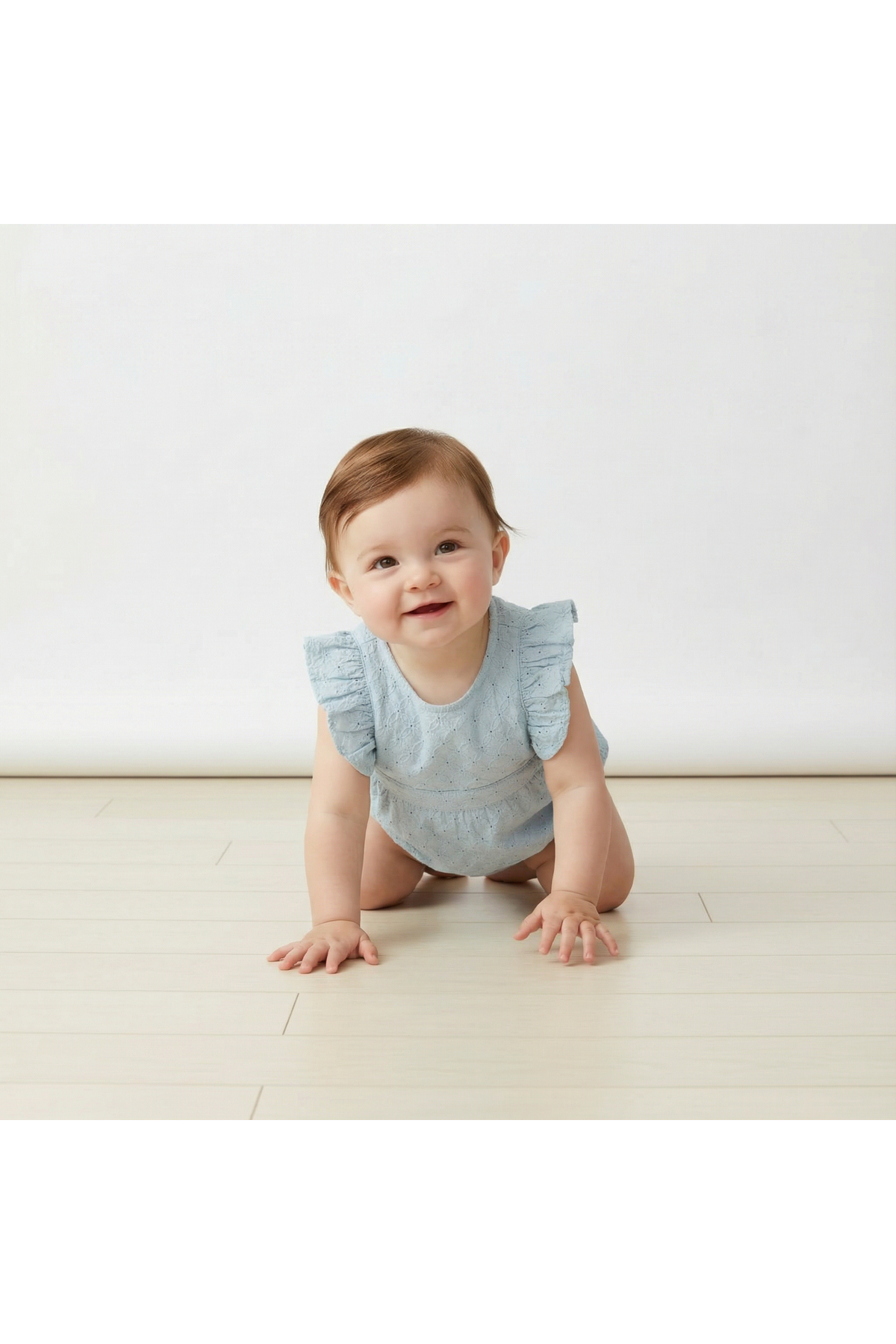 A smiling baby crawls on a light wooden floor, wearing a light blue organic cotton romper with eyelet detailing and flutter sleeves. The simple background emphasizes the outfits delicate design.