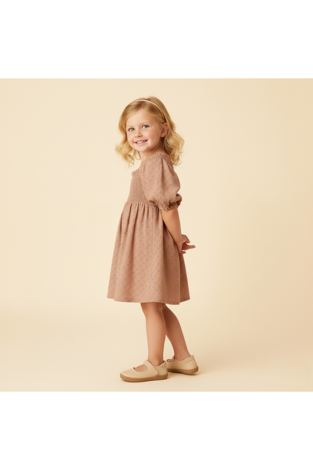 A young girl models a clay-colored organic cotton eyelet bubble sleeve dress, featuring delicate embroidery. She stands against a soft, neutral background, smiling and wearing beige shoes.