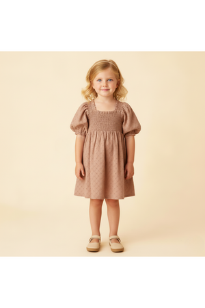 A young girl stands wearing a clay-colored organic cotton dress with bubble sleeves and delicate eyelet detailing. The background is a soft, neutral tone, enhancing the dresss gentle appearance.