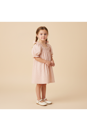 A young girl stands in profile, wearing a blush pink organic cotton dress with eyelet details and bubble sleeves. The soft fabric complements her playful style against a neutral background.
