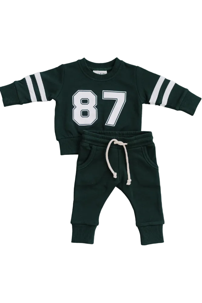 A dark green French terry cotton two-piece set featuring a crewneck sweatshirt with white stripes and a number 87 print, paired with matching pants that have an elastic waistband.