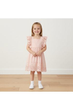 A young girl stands smiling in a blush pink organic cotton dress featuring delicate eyelet detailing and flutter sleeves. The dress is perfect for playdates and casual outings.