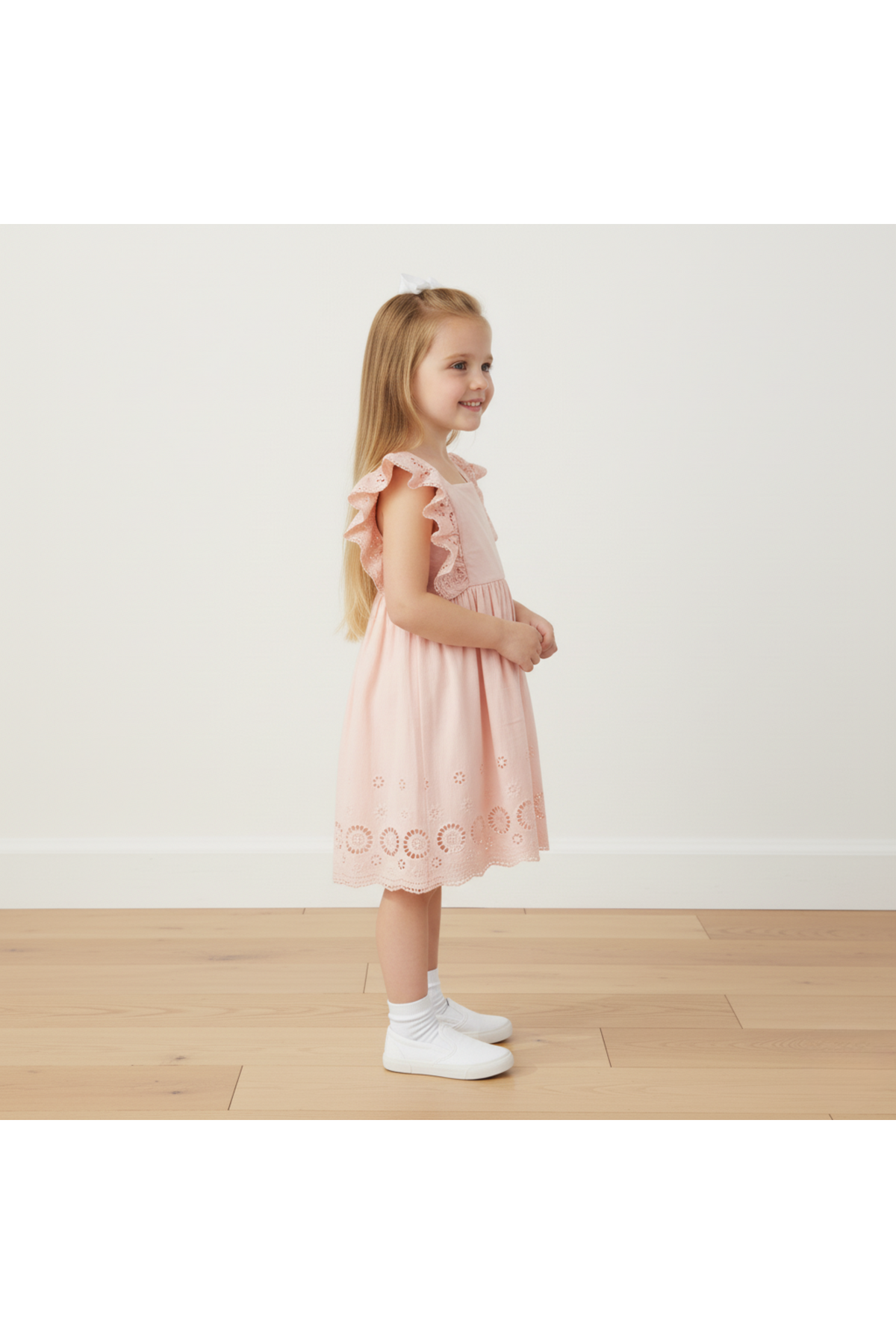 A young girl stands sideways, wearing a blush organic cotton dress with delicate flutter sleeves and eyelet details.