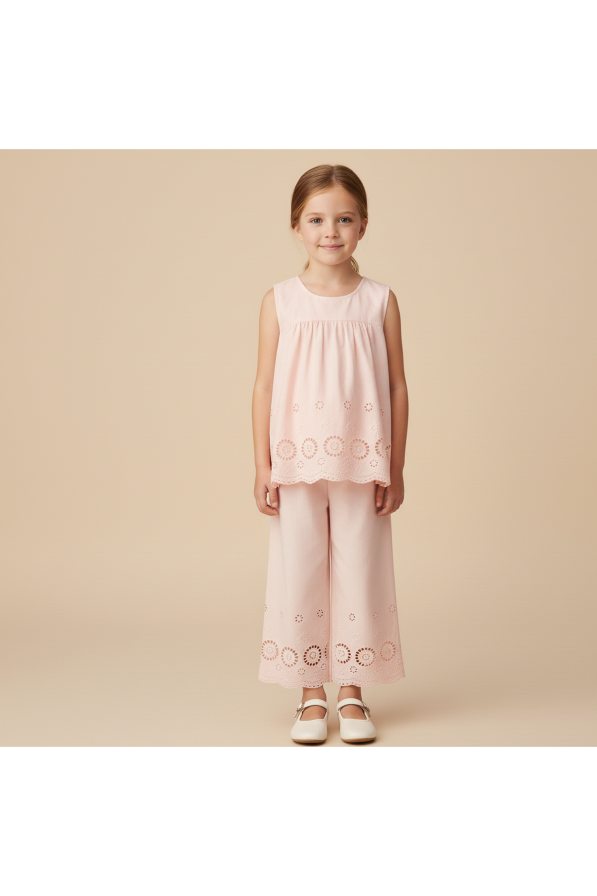 A young girl stands wearing a blush organic cotton eyelet top and matching pant set, featuring delicate embroidered details. The outfit is perfect for warm-weather outings and celebrations.